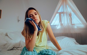 Woman sitting in bed with eyes closed holding cold compress to face