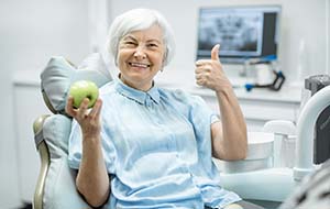 smiling older woman in the dental chair holding an apple and giving a thumbs-up
