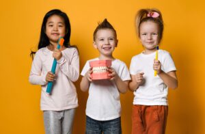 Three smiling children in white shirts holding large models of dental gear in front of an orange background