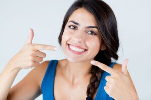 Woman with brown hair in blue shirt smiling and pointing to her teeth with her pointer fingers